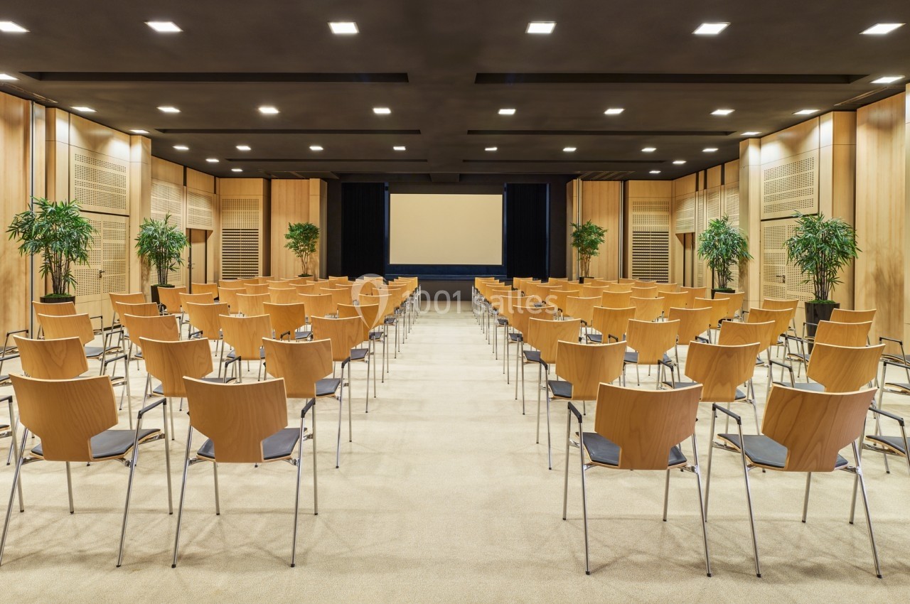 Salle de conférence moderne avec rangées de chaises en bois, écran de projection et plantes décoratives.