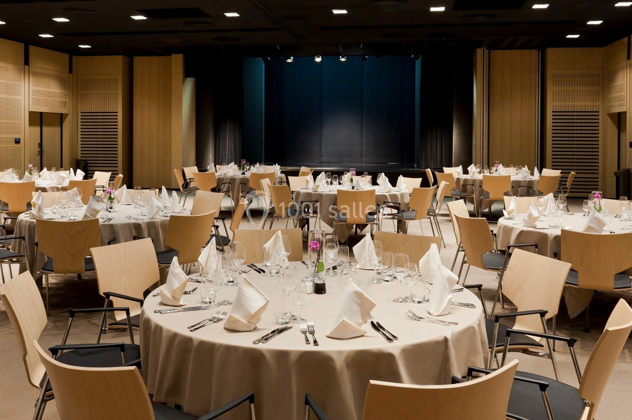 Salle de réception avec tables rondes dressées, nappes beiges, serviettes pliées et chaises en bois clair.