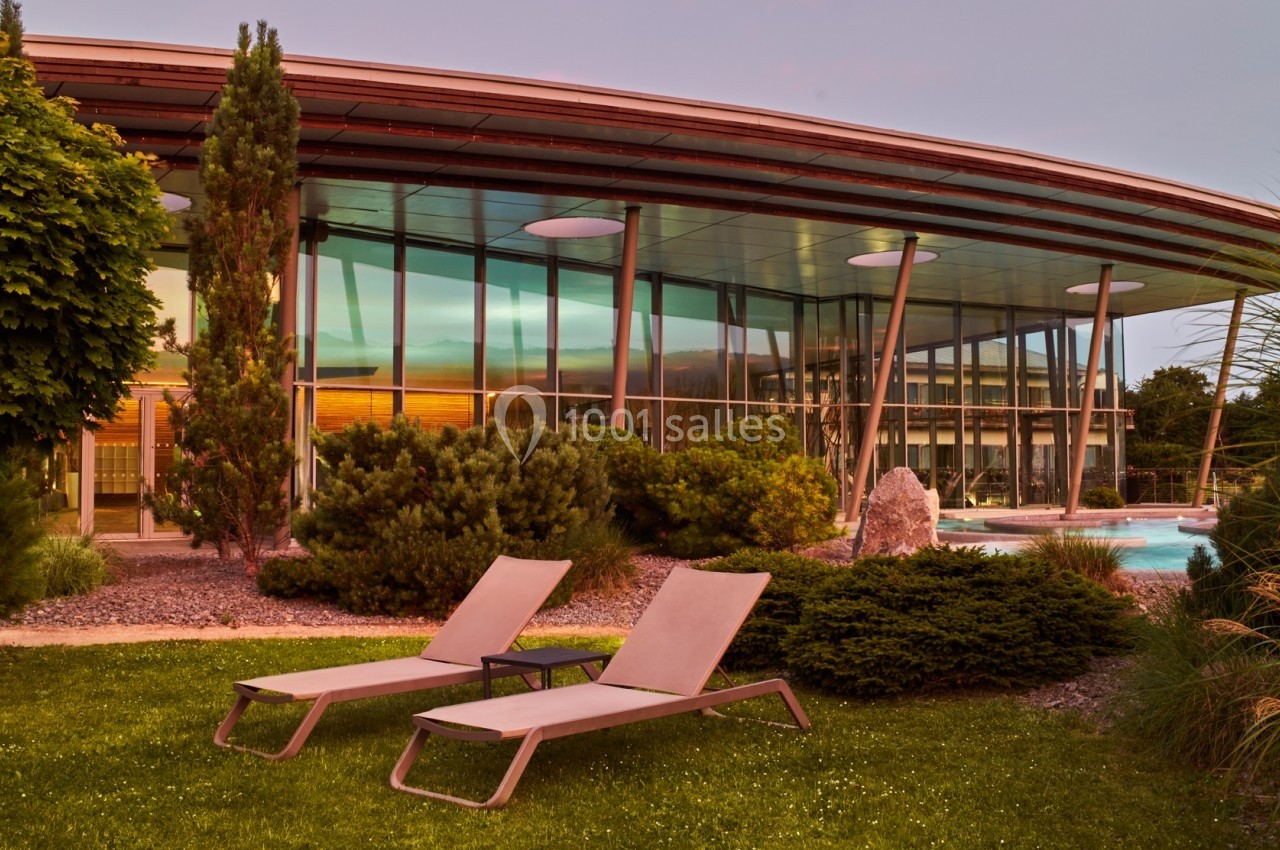 Façade vitrée d'un bâtiment moderne entouré de végétation, avec deux chaises longues sur une pelouse au premier plan.