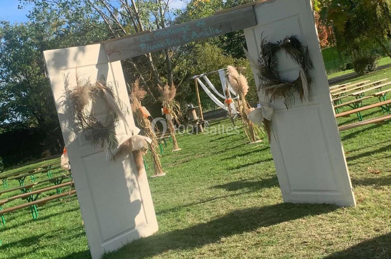 Deux portes blanches décorées de couronnes végétales encadrent une allée menant à une cérémonie en plein air.