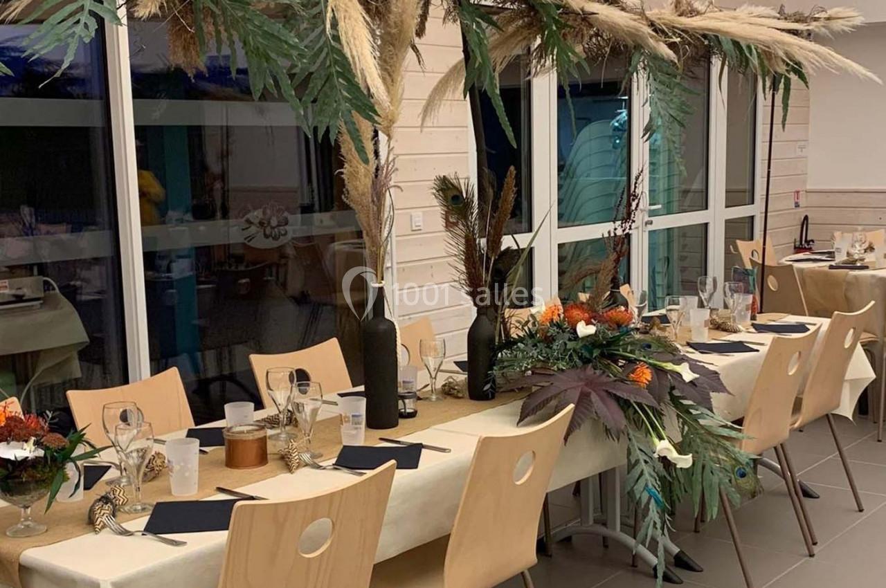 Salle décorée pour un repas, avec des tables dressées, ornées de compositions florales et de feuillages suspendus.