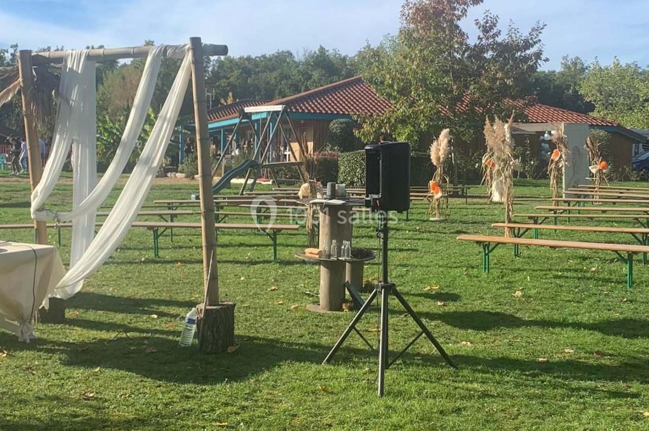 Espace extérieur avec bancs en bois, arche décorée de voilages blancs et pelouse sous un ciel dégagé.