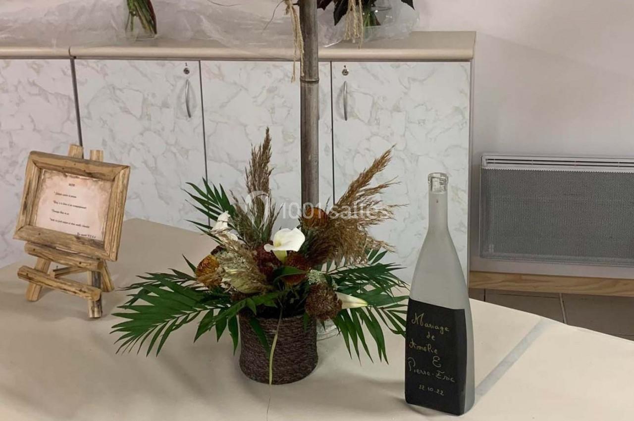 Arrangement floral avec des feuilles et des fleurs séchées sur une table, accompagné d'une bouteille personnalisée.