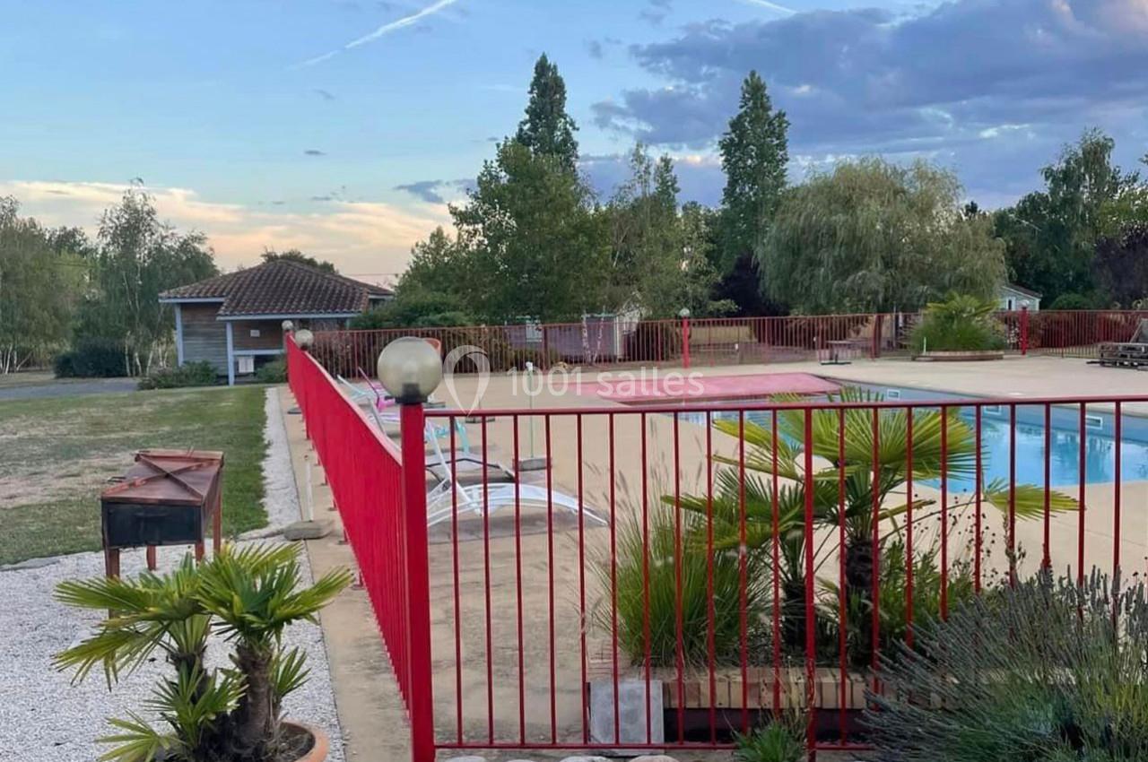 Clôture rouge entourant une piscine extérieure avec transats, arbres et ciel nuageux en arrière-plan.