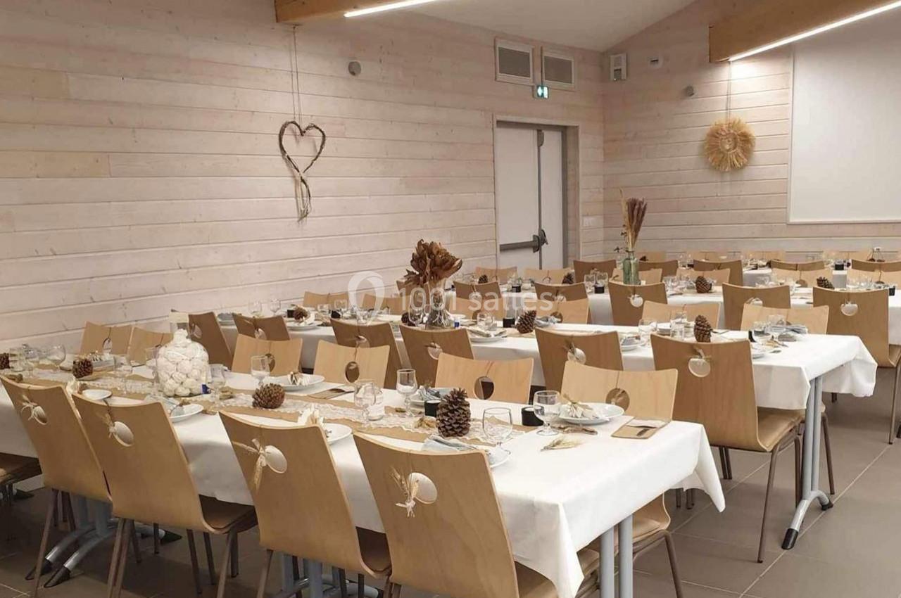 Salle décorée pour un repas, avec des tables alignées, nappes blanches, décorations naturelles et chaises en bois.