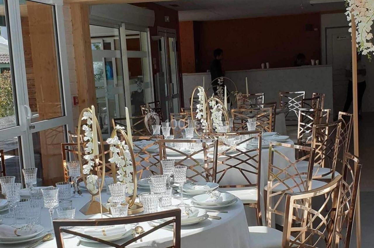 Salle lumineuse avec tables rondes dressées, décorées de fleurs blanches et bougies, prête pour un événement.