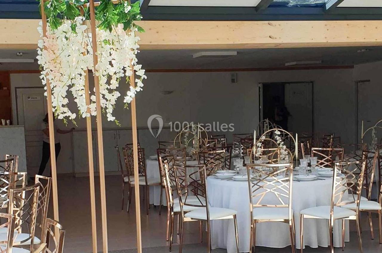 Salle de réception avec tables rondes dressées, chaises dorées et décoration florale suspendue.
