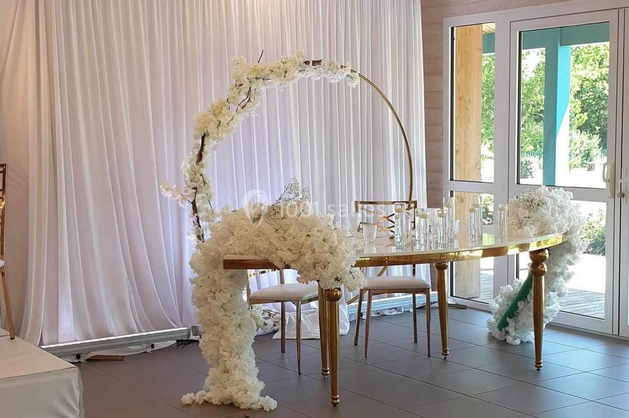 Table décorée pour un événement, ornée de fleurs blanches et d'une arche circulaire, devant un rideau blanc.