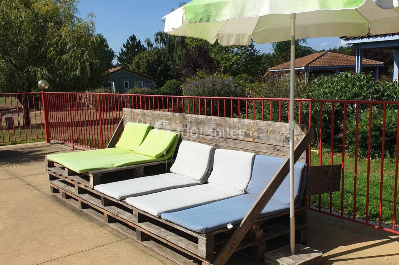 Banc en palettes avec coussins colorés et parasol, installé sur une terrasse ensoleillée près d'une barrière rouge.