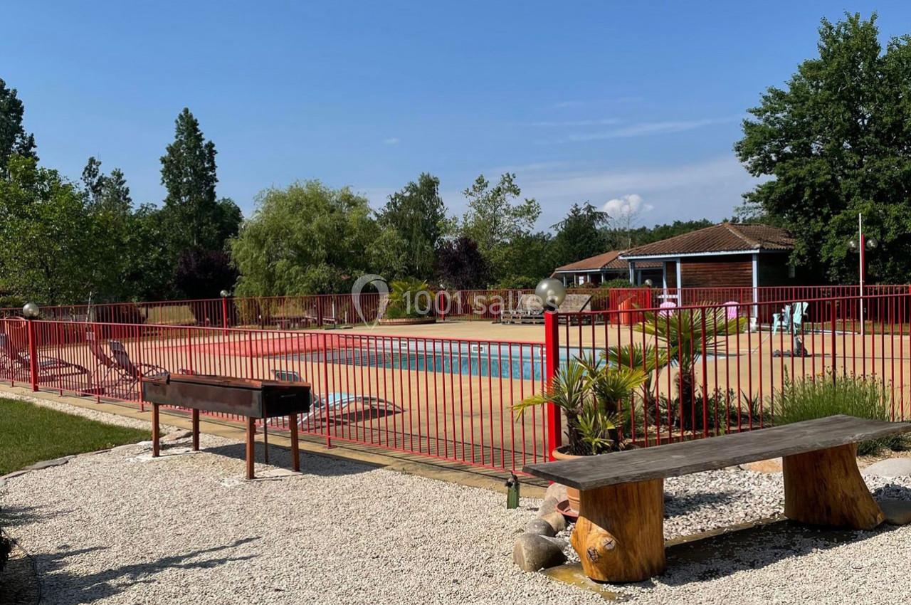 Piscine extérieure entourée d'une barrière rouge, avec un banc en bois et un barbecue au premier plan.