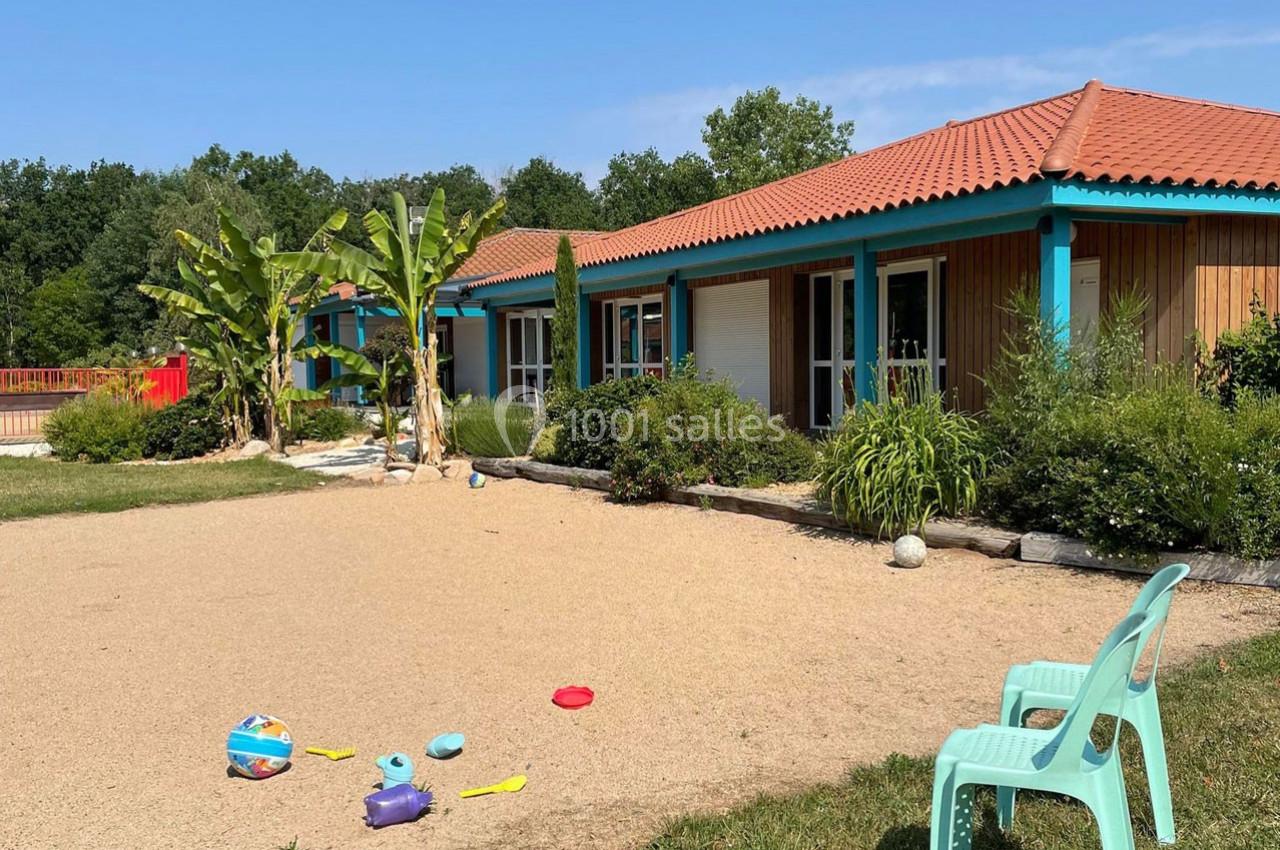 Cour extérieure d'une maison avec terrasse, plantes tropicales, jouets pour enfants au sol et chaises en plastique.