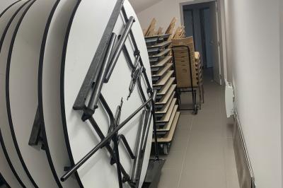 Tables rondes empilées verticalement et chaises empilées dans un couloir étroit avec un sol en carrelage gris.