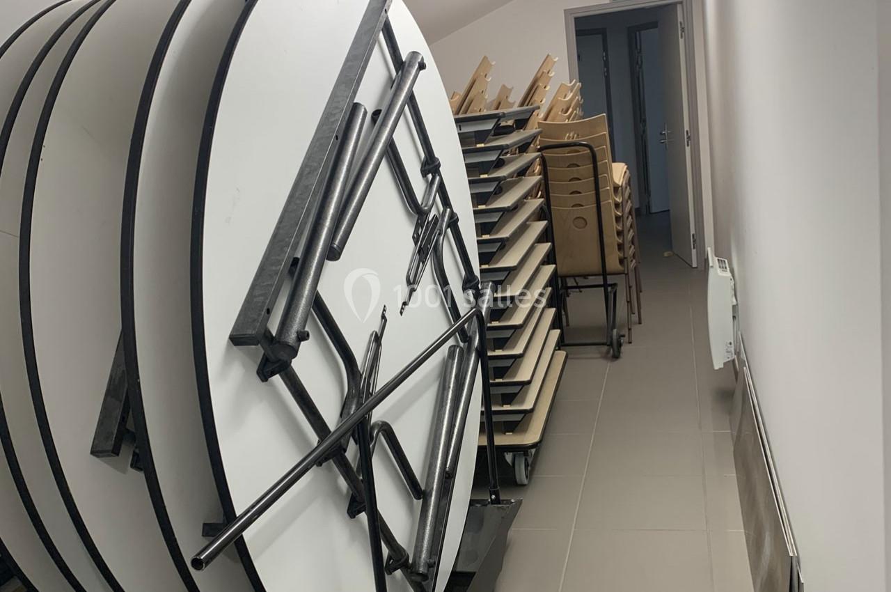 Tables rondes empilées verticalement et chaises empilées dans un couloir étroit avec un sol en carrelage gris.