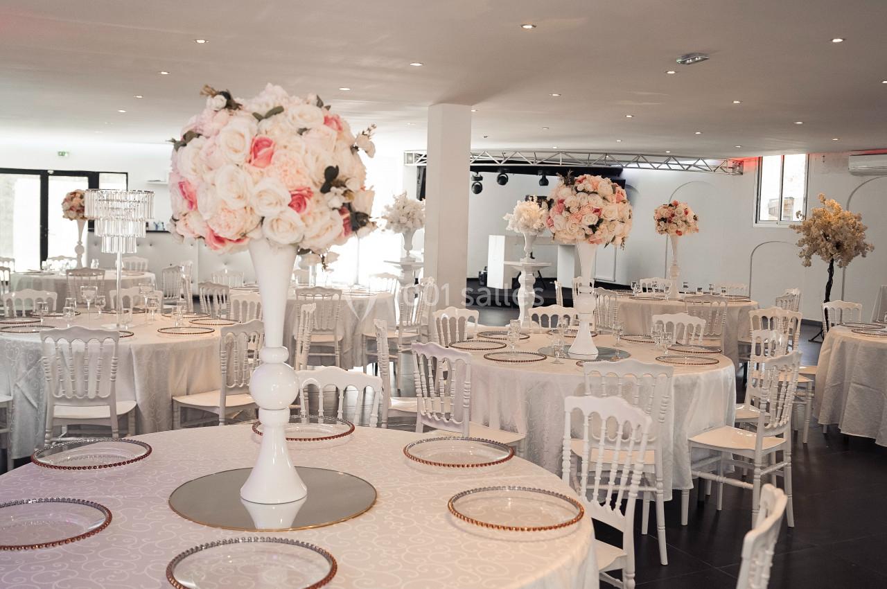 Salle de réception décorée avec des tables rondes, nappes blanches, chaises assorties et centres de table fleuris.