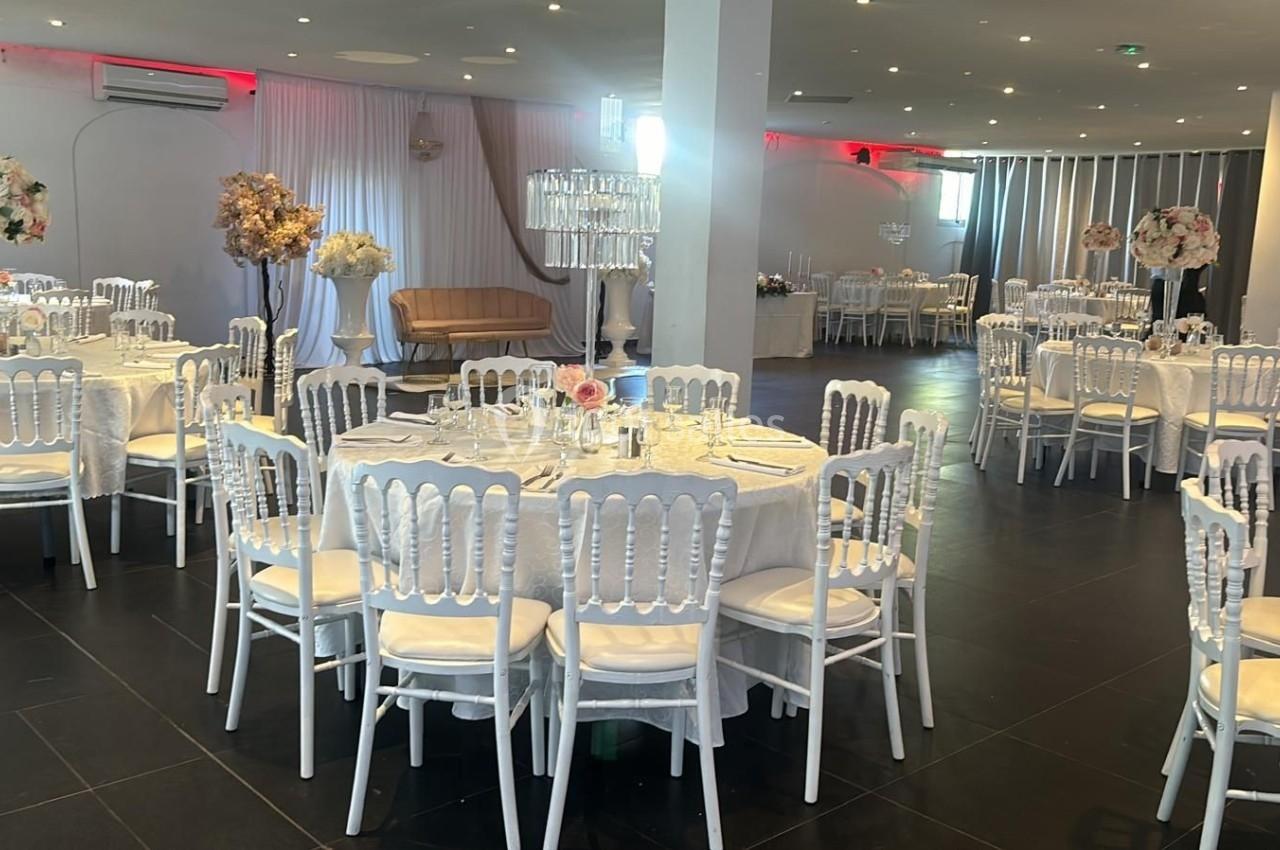 Salle de réception élégante avec tables rondes dressées, chaises blanches et décorations florales.