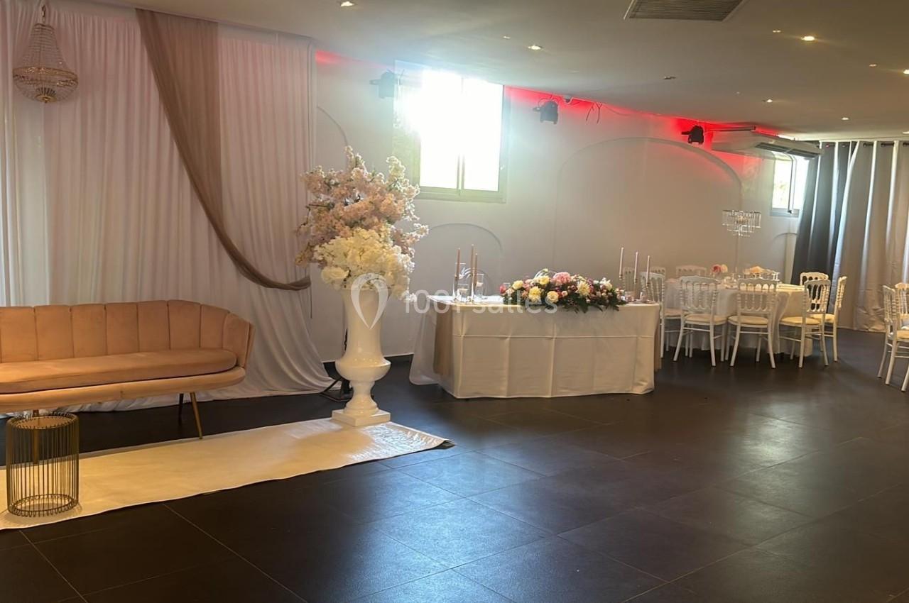 Salle décorée pour un événement avec canapé beige, rideaux blancs, fleurs et tables dressées.