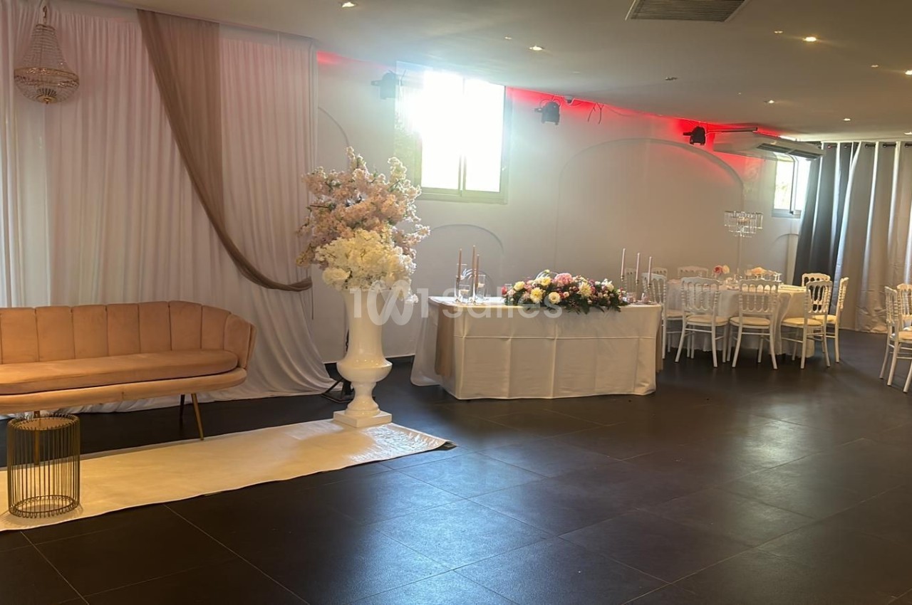 Salle décorée pour un événement avec canapé beige, rideaux blancs, fleurs et tables dressées.