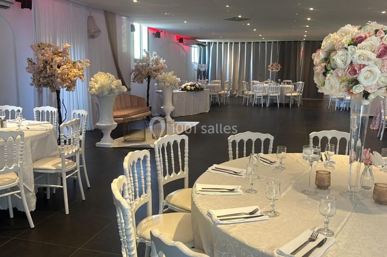 Salle de réception élégamment décorée avec des tables rondes, nappes blanches, chaises blanches et arrangements floraux.