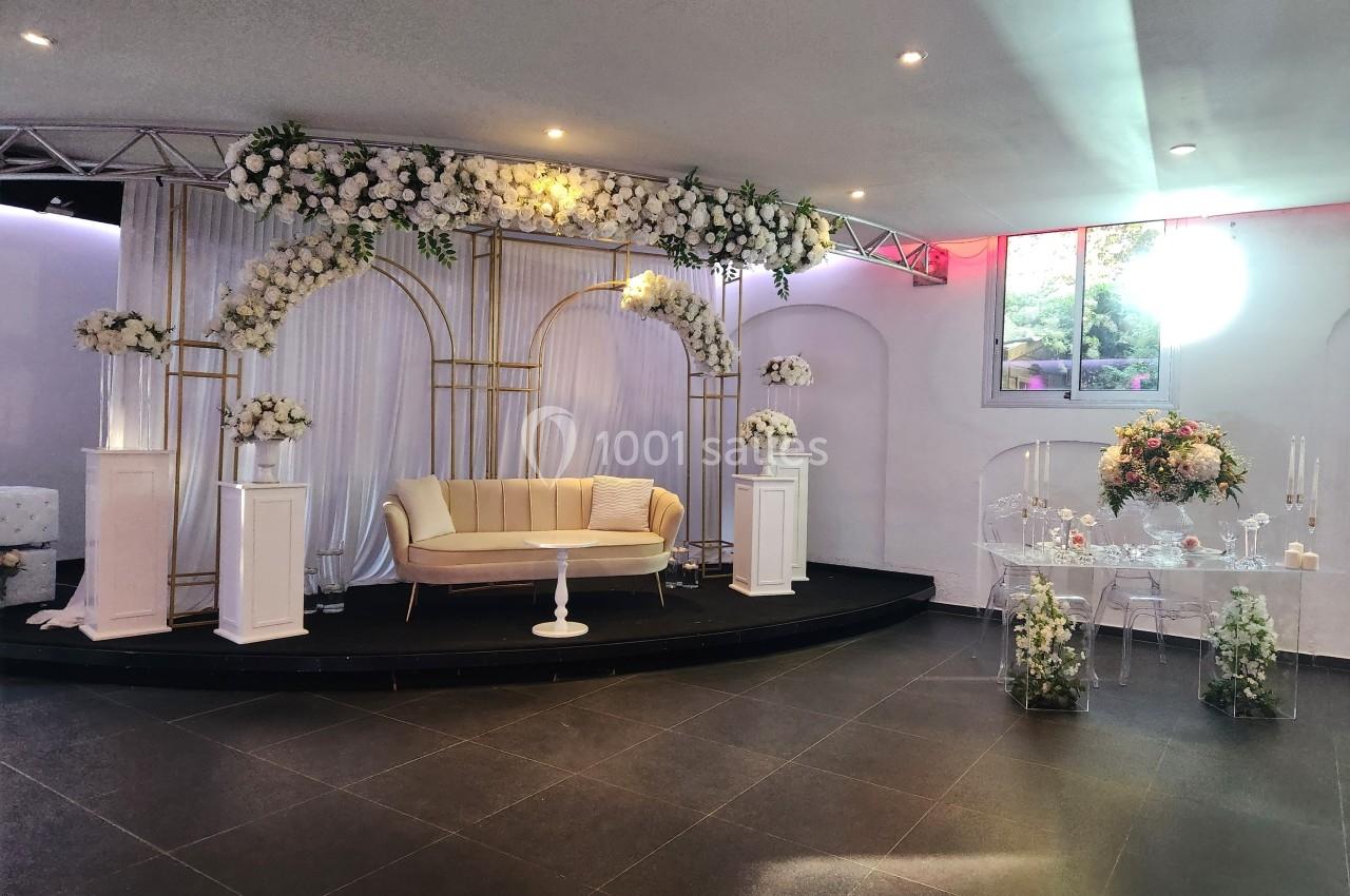 Décoration de salle avec arche florale blanche, canapé beige, table transparente et éclairage naturel.