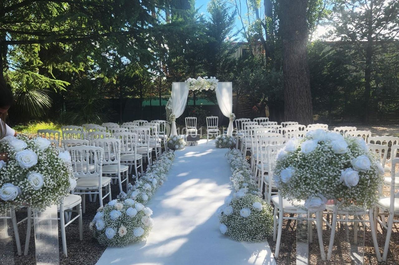 Allée centrale décorée de fleurs blanches menant à une arche nuptiale dans un cadre extérieur boisé.