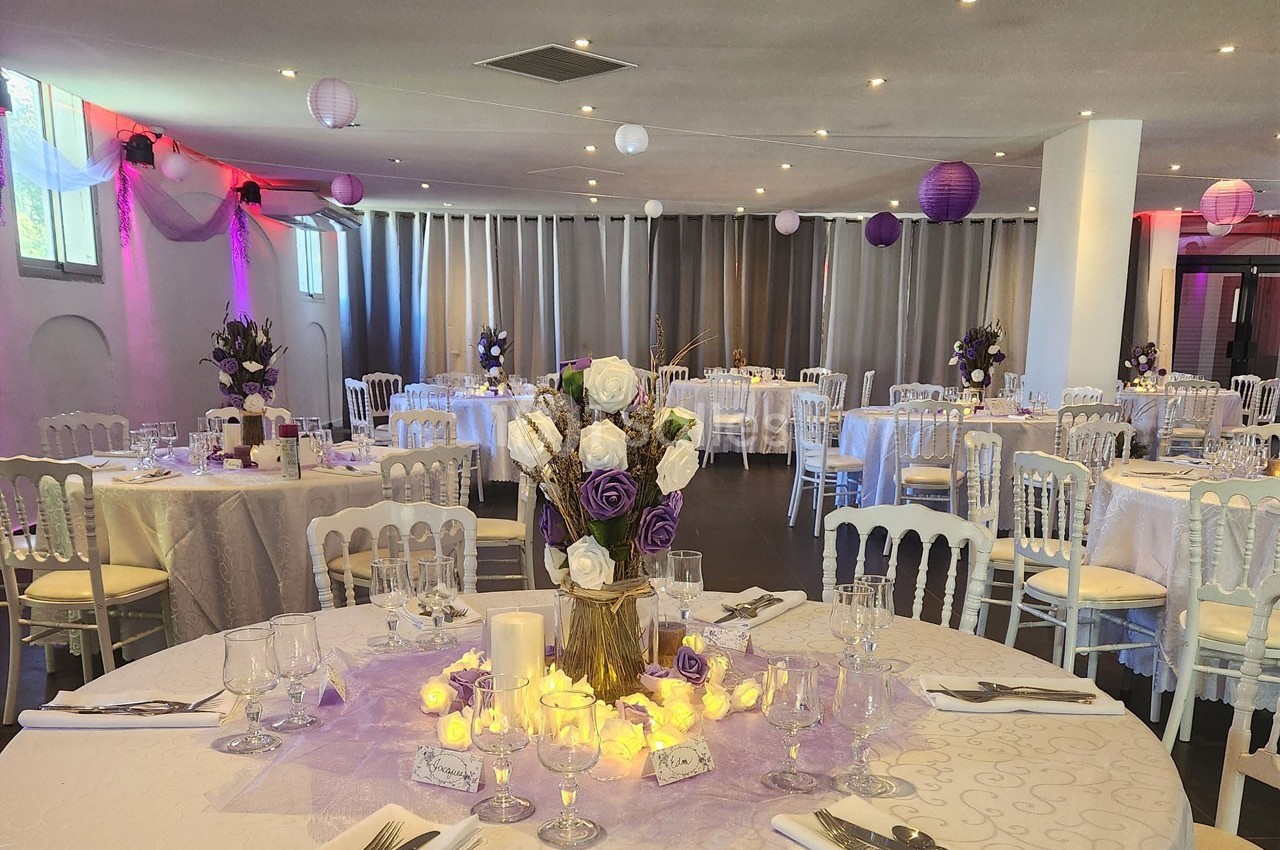 Salle de réception décorée avec des tables rondes, nappes blanches, fleurs violettes et éclairage tamisé.