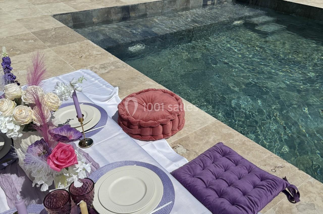 Table de pique-nique élégante avec coussins colorés et vaisselle raffinée, disposée au bord d'une piscine ensoleillée.