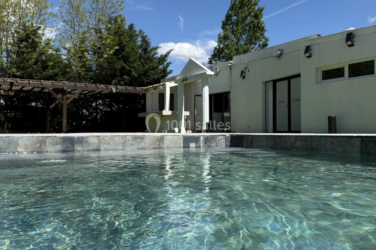 Piscine extérieure avec eau claire, entourée d'une terrasse, d'arbres et d'un bâtiment moderne sous un ciel dégagé.