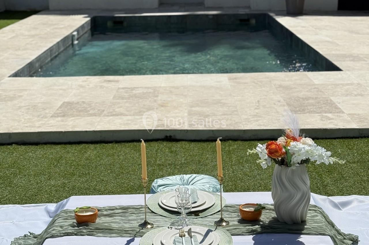 Table dressée avec vaisselle élégante, bougies et fleurs, disposée devant une piscine entourée de pierre et d'herbe.