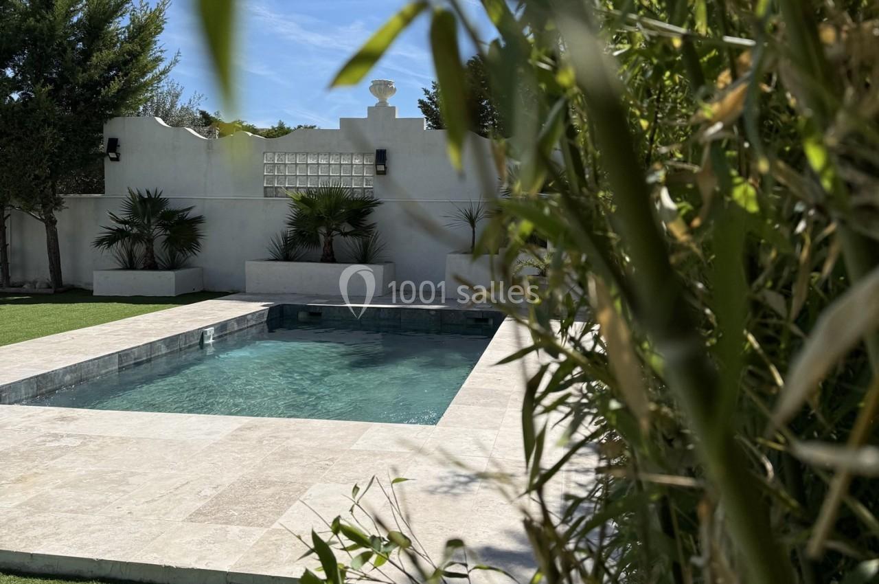Piscine rectangulaire entourée de végétation et de dalles claires, avec un mur blanc en arrière-plan.