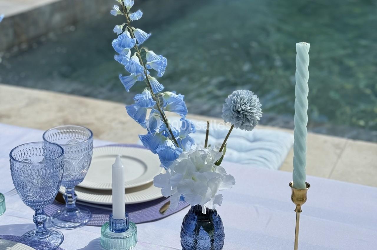 Table dressée près d'une piscine avec des fleurs bleues et blanches, des bougies et de la vaisselle en verre.