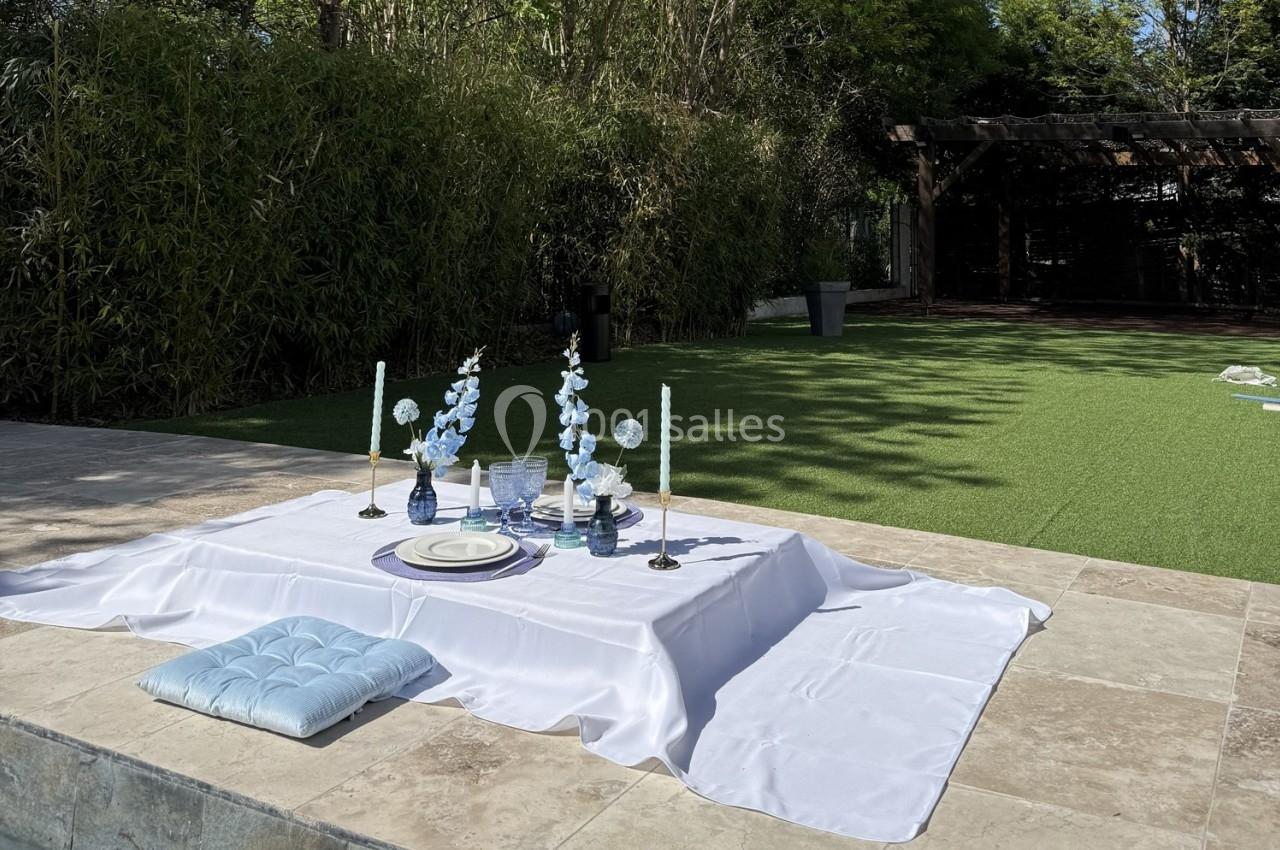 Table basse dressée sur une nappe blanche avec des bougies et des fleurs bleues, située en extérieur près d'une pelouse.