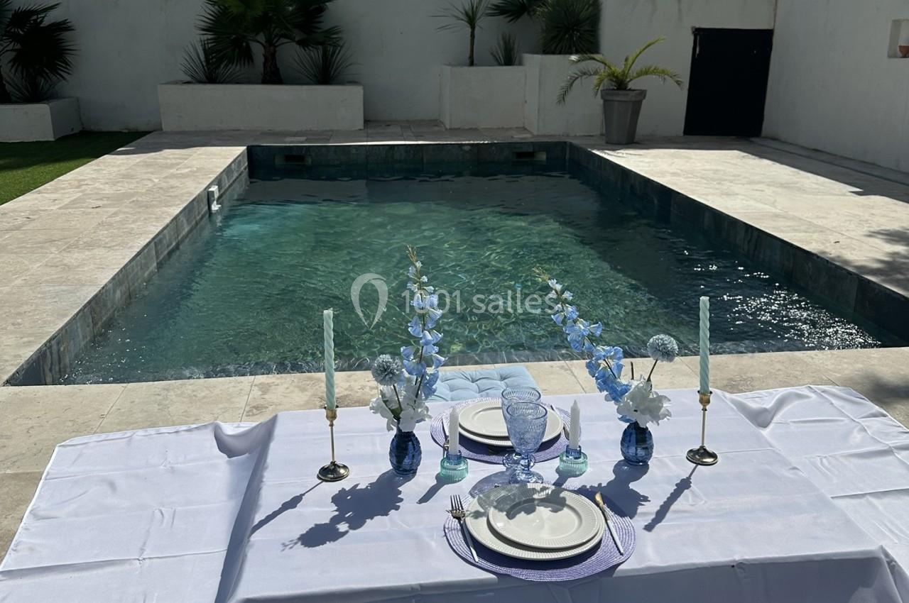 Table dressée avec vaisselle et fleurs bleues près d'une piscine entourée de dalles claires et de végétation.