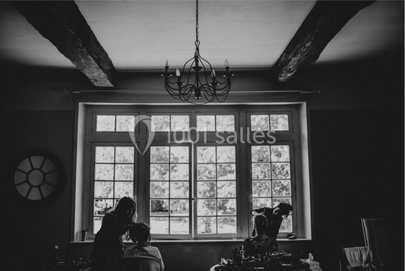 Deux personnes dans une pièce sombre avec une grande fenêtre, éclairées par la lumière extérieure, sous un lustre.