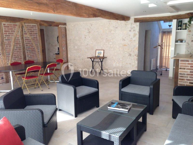 Salle lumineuse avec murs en pierre, poutres apparentes, mobilier en rotin noir et table avec chaises rouges pliantes.