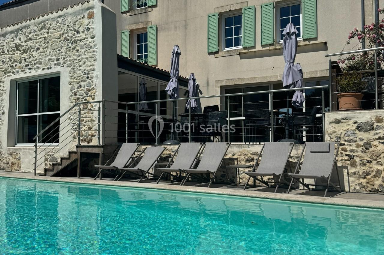 Chaises longues alignées au bord d'une piscine devant une maison en pierre avec volets verts et parasols fermés.