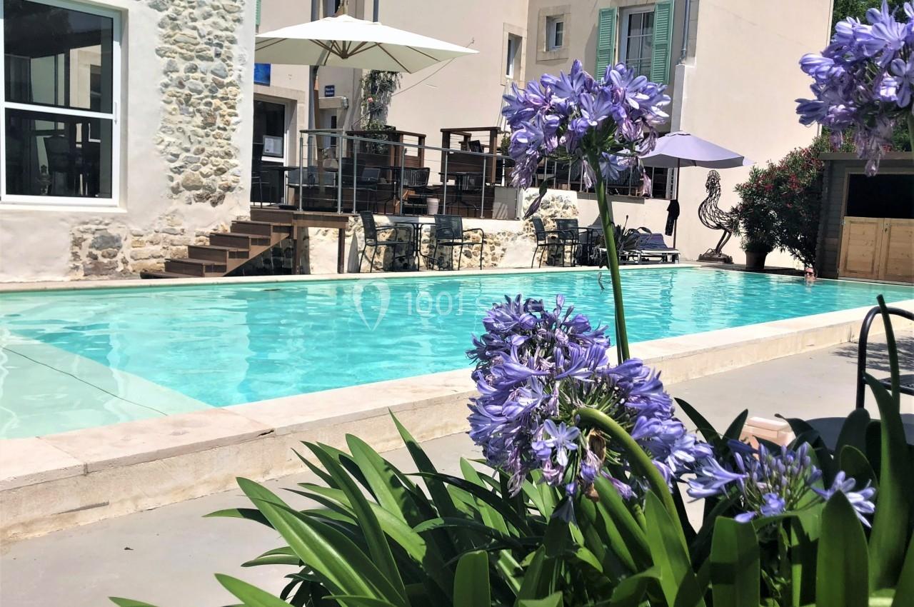 Piscine extérieure bordée de fleurs violettes, avec terrasse ombragée et bâtiments en arrière-plan.