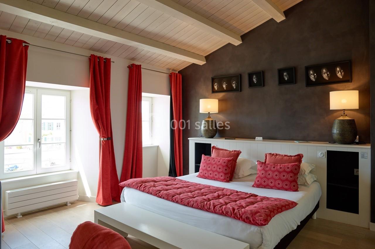 Chambre lumineuse avec lit double, murs sombres, poutres apparentes, rideaux rouges et décoration moderne.