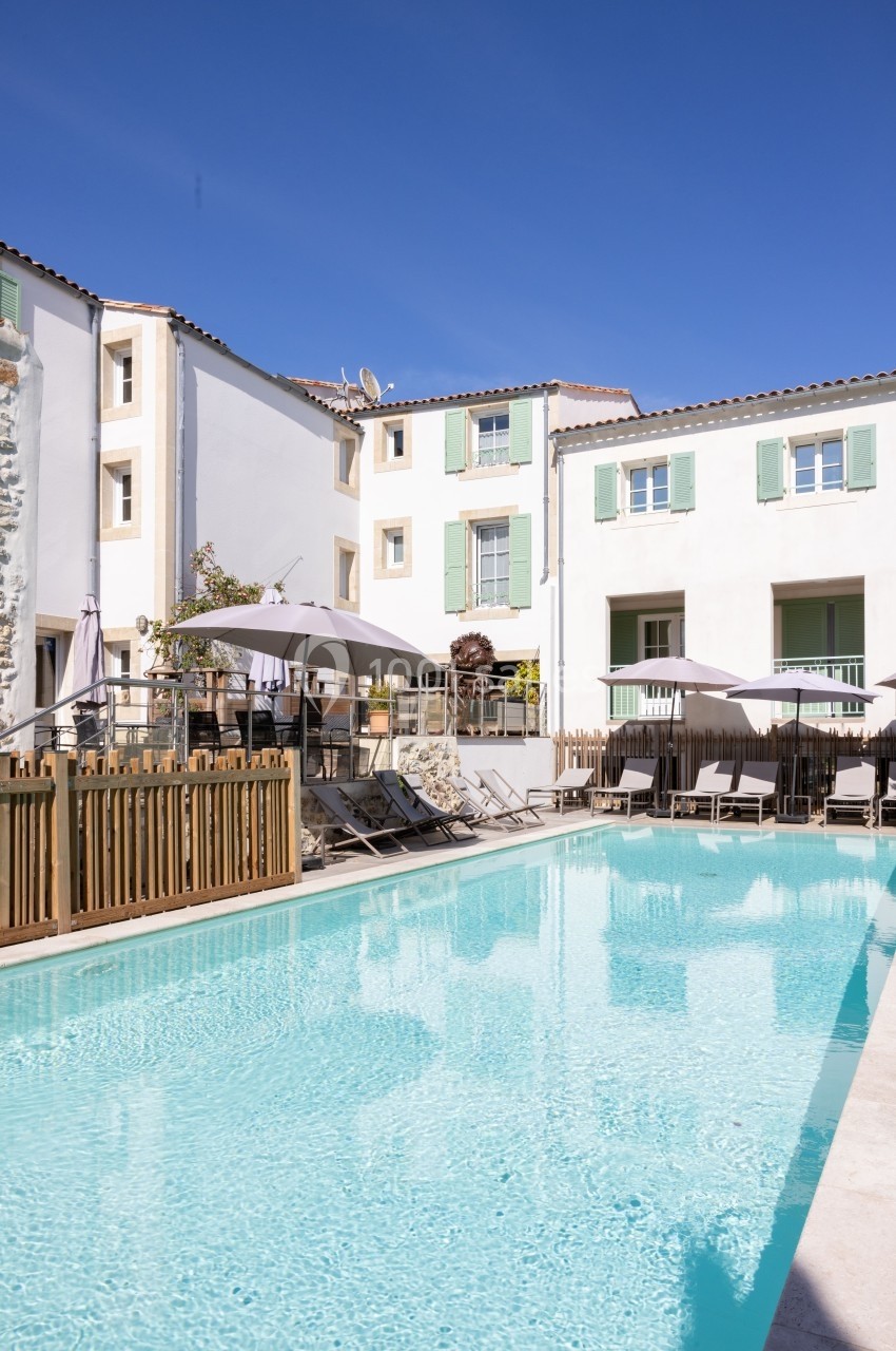 Piscine extérieure entourée de transats et parasols, avec des bâtiments aux volets verts en arrière-plan.