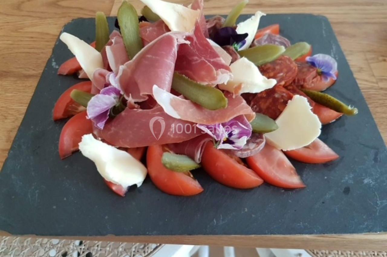 Assiette de charcuterie avec jambon, cornichons, fromage, tranches de tomate et fleurs comestibles sur ardoise noire.
