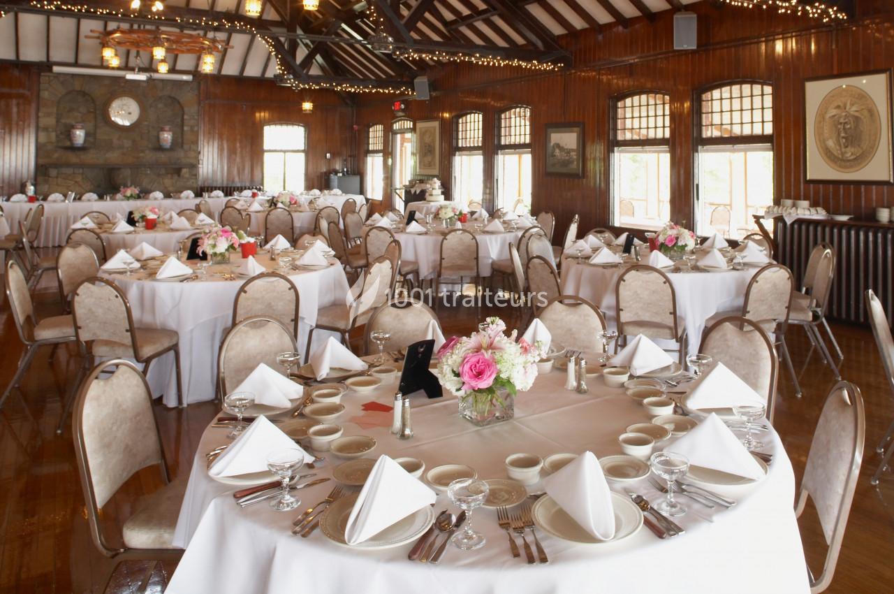Salle de réception avec tables rondes dressées, nappes blanches, serviettes pliées et centres de table fleuris.