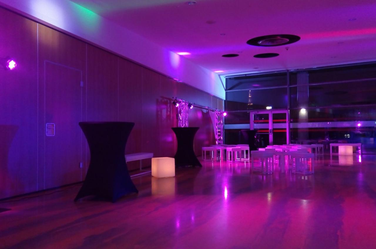 Salle de réception moderne avec éclairage violet, tables hautes noires et mobilier blanc, vue nocturne sur l'extérieur.