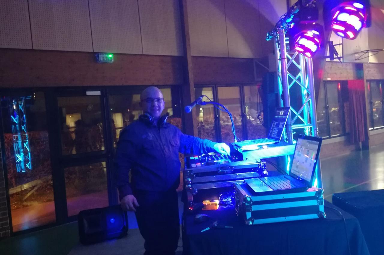 Un DJ souriant se tient derrière une table de mixage éclairée par des lumières colorées dans une salle de réception.