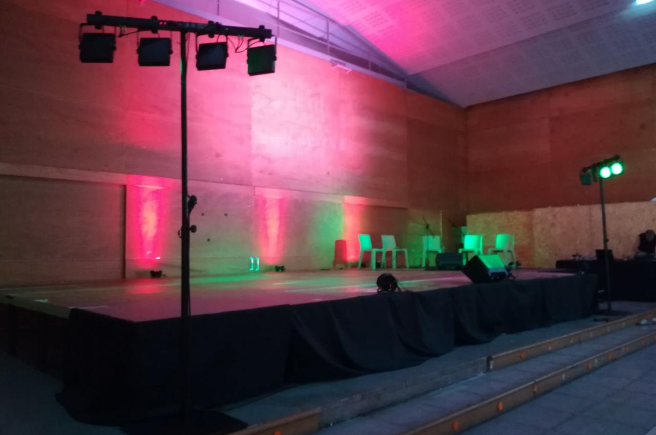 Scène vide avec éclairage coloré rouge et vert dans une salle en bois, chaises blanches en arrière-plan.