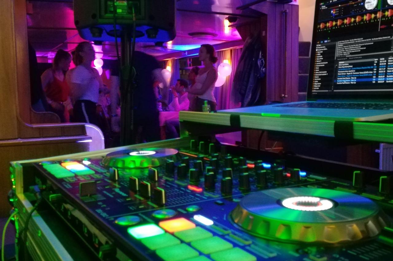 Console de mixage éclairée au premier plan, avec des invités dansant et discutant dans une ambiance festive en arrière-plan.