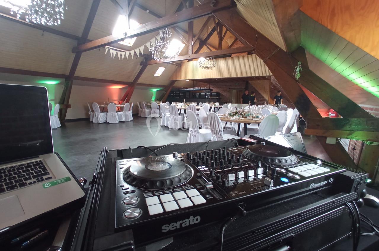 Console de DJ en premier plan dans une salle de réception décorée avec des tables dressées et des lumières colorées.