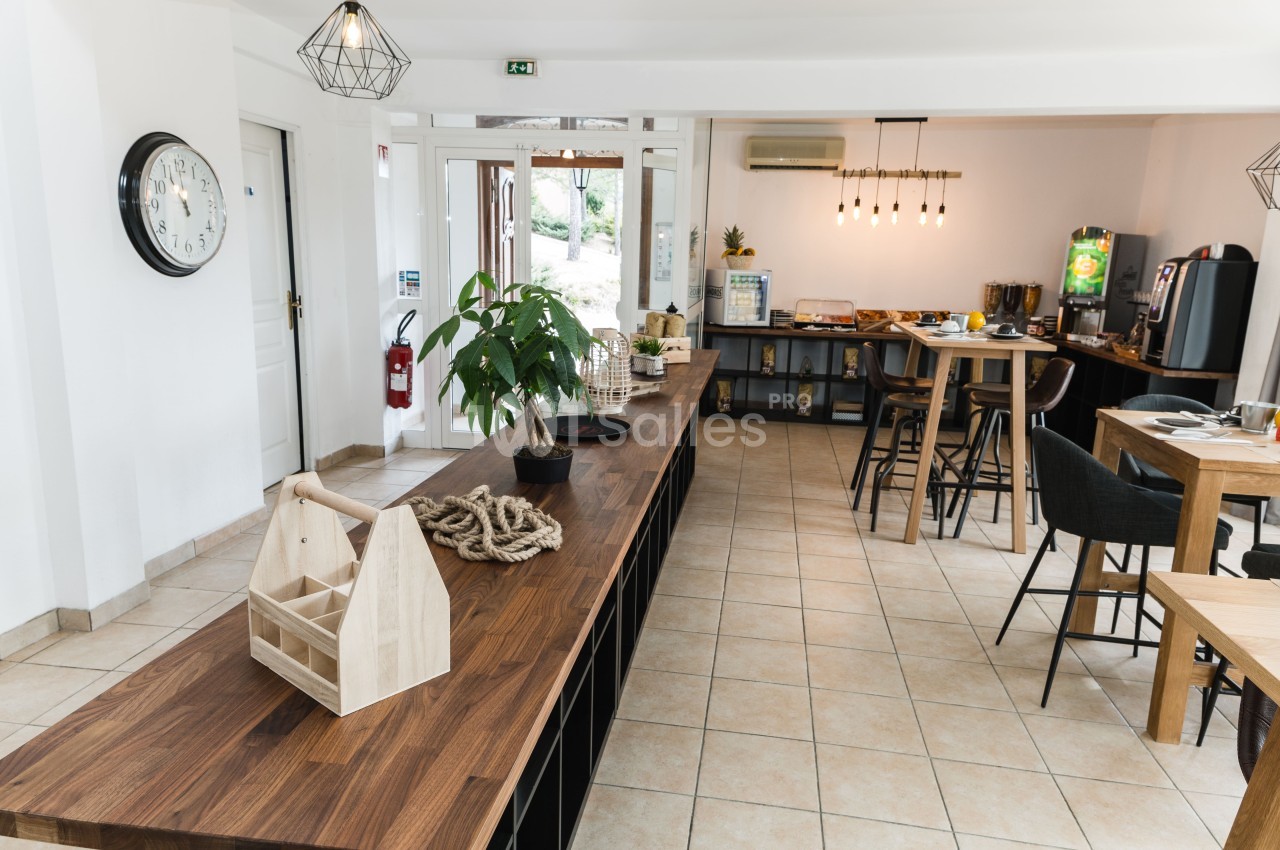 Salle de petit-déjeuner aux carrelages clairs, avec un long plan de travail en bois foncé, des tables hautes et des tables en bois aux chaises sombres, et un buffet au fond.