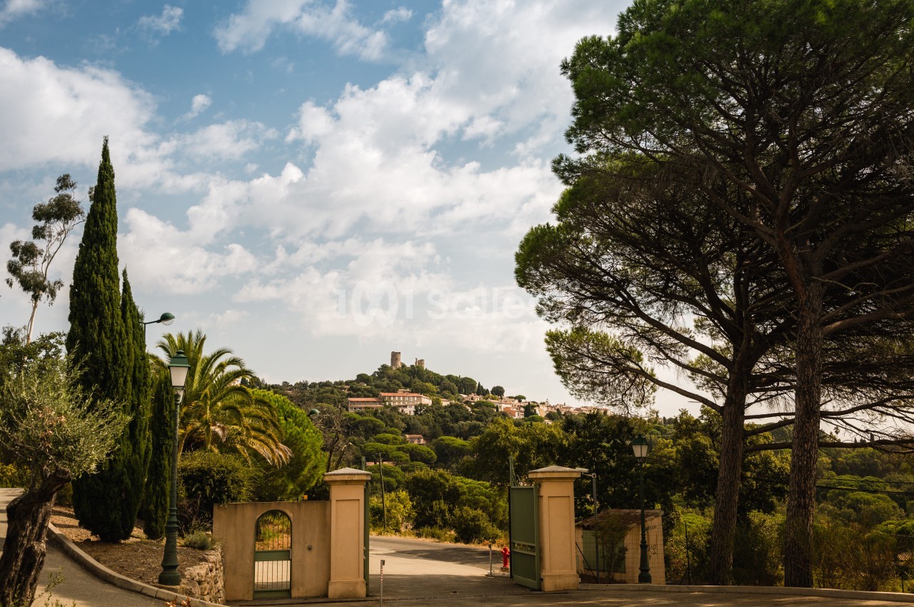 Entrée d'une propriété en hauteur avec portail vert et piliers crème, bordée de cyprès, palmiers et pins parasols, avec un village perché au loin sous un ciel nuageux.