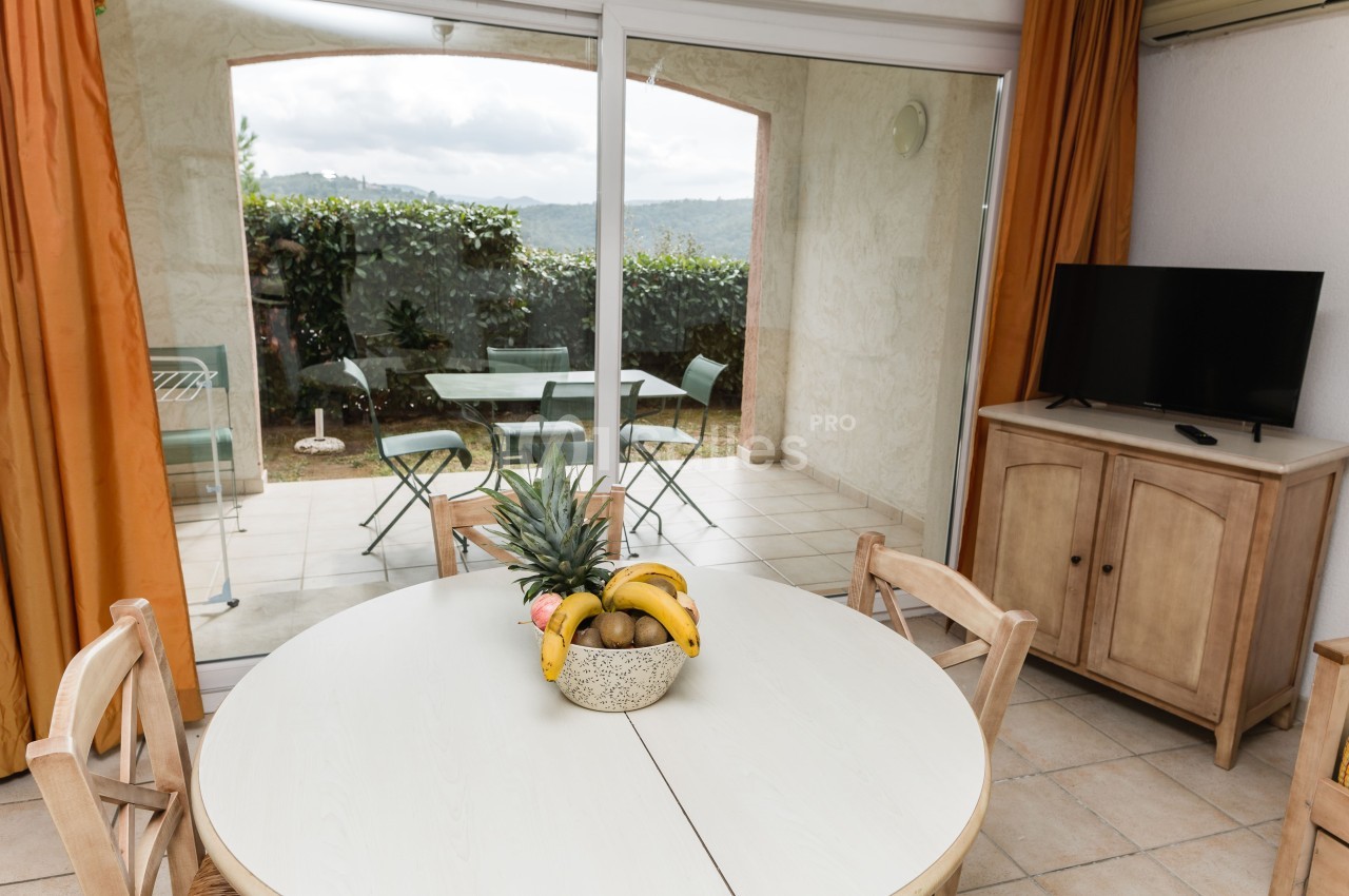 Table ronde blanche dressée d'une corbeille de fruits (ananas, bananes, kiwis) dans une pièce lumineuse, avec vue sur une terrasse carrelée et un jardin par la baie vitrée.