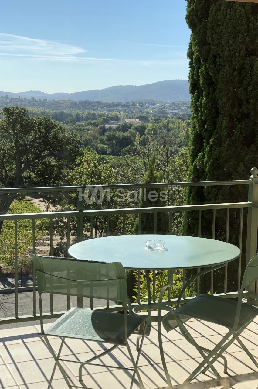 Balcon carrelé avec une table ronde et deux chaises en métal vert, face à un paysage de collines boisées et de cyprès sous un ciel bleu.