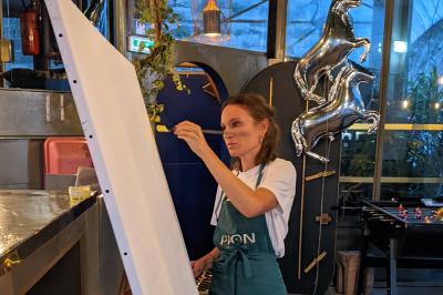 Une femme peint sur une grande toile blanche dans un espace intérieur décoré de plantes et de ballons argentés.